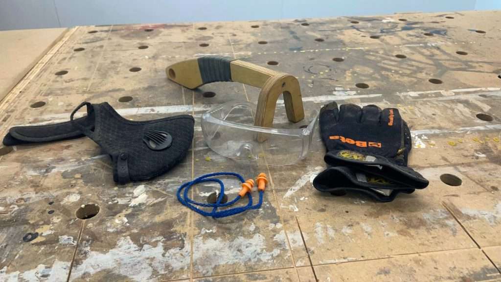 Personal protective equipment laid out in a safe workshop: gloves, earplugs, safety glasses, dust mask, and push stick.