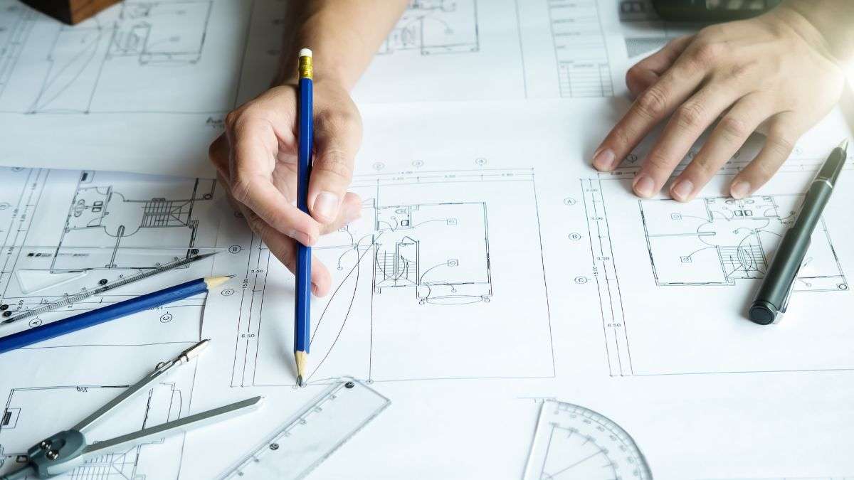 Close-up of hands sketching workshop plans on paper with pencils and rulers, representing the early stages of planning a woodworking shop.